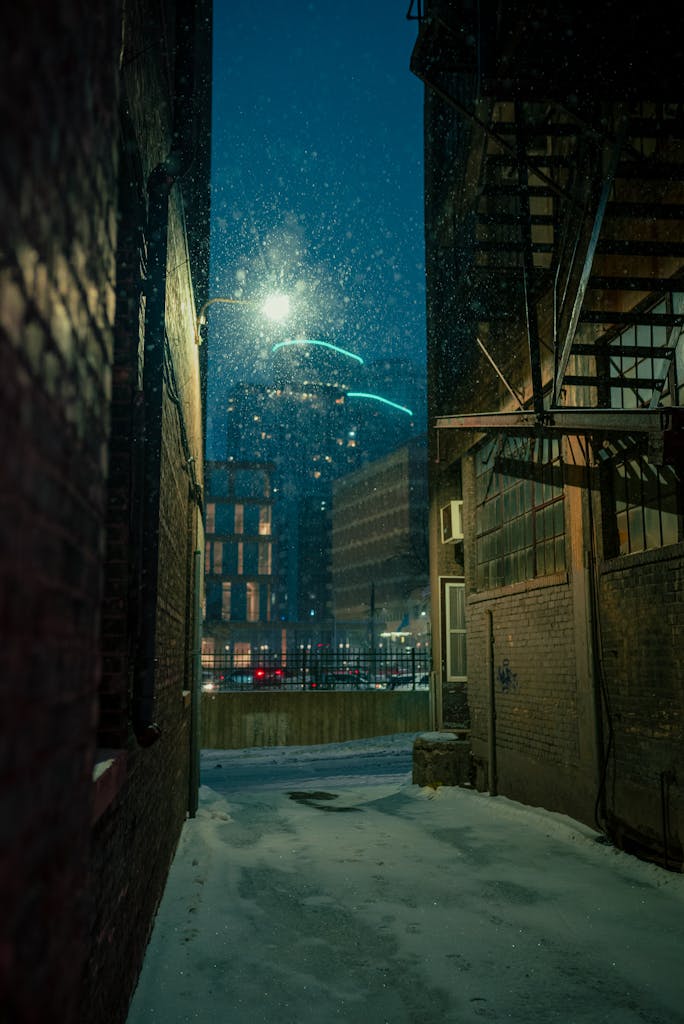 Snow falls quietly in a dimly lit urban alley, with glowing city skyscrapers in the background.
